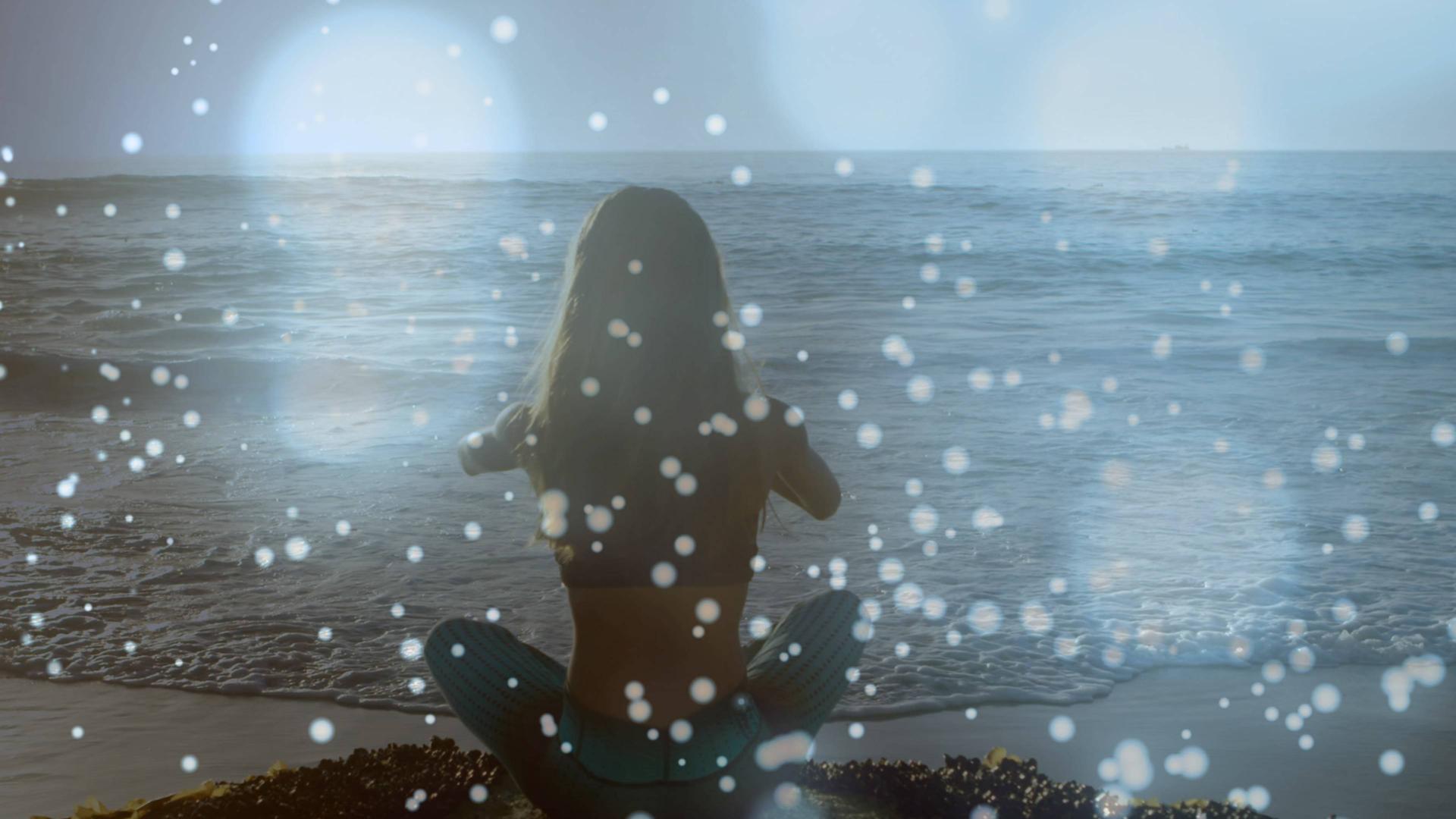 Eine Person in Yogapose sitzt dem Betrachter mit dem Rücke zugewandt am Meer. Über dem Bild liegt eine Fläche aus mystischem Schillern, was dem Bild eine spirituelle Stimmung verleiht.